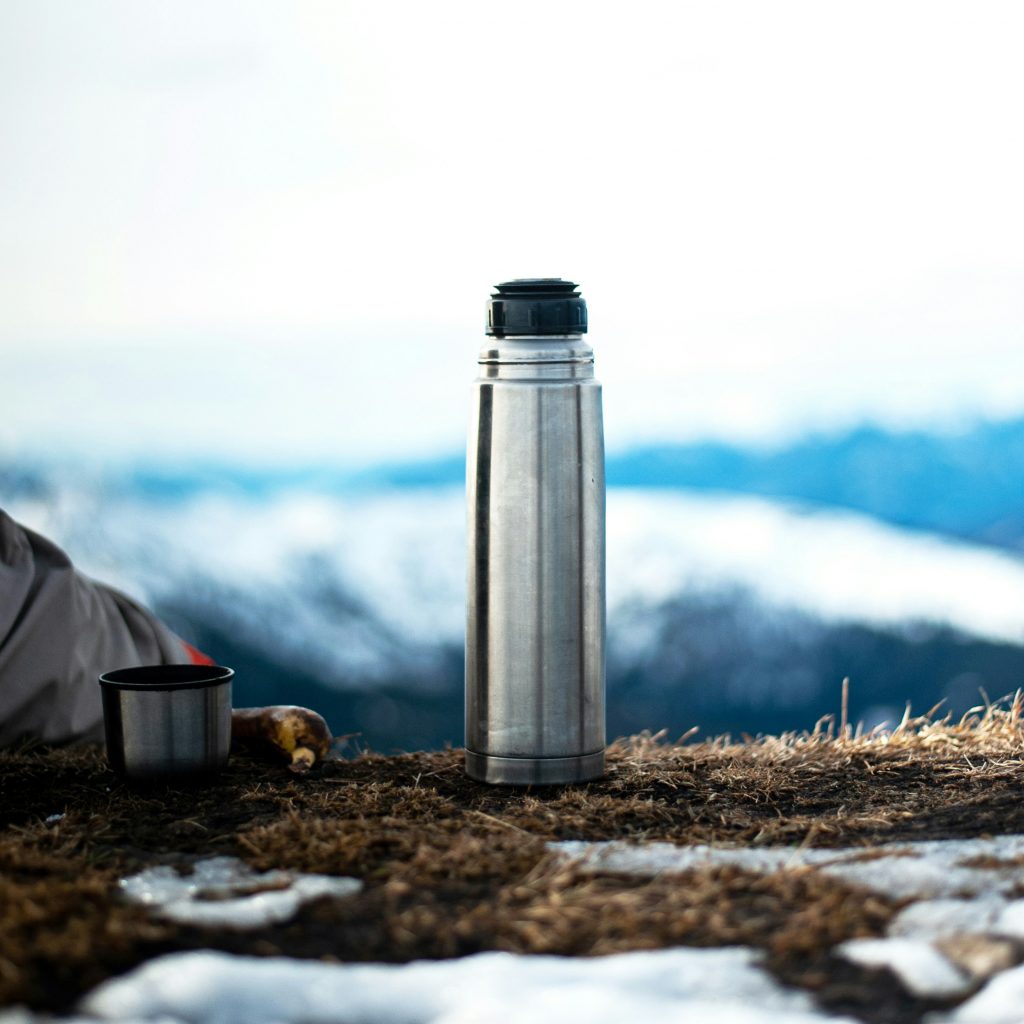 Thermoskanne vor winterlicher unscharfer Aussicht, steht auf braunem Gras mit Schneeflecken, daneben der Deckel. 
