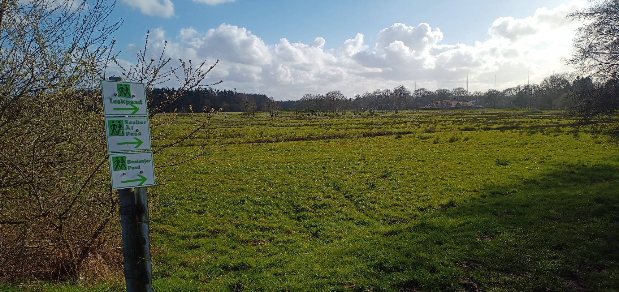 Blick auf Weiden, im Hintergrund Wald, oben blauer Himmel mit weißen Wolken, im Vordergrund ein Pfahl mit Hinweisschildern auf verschiedene Paads